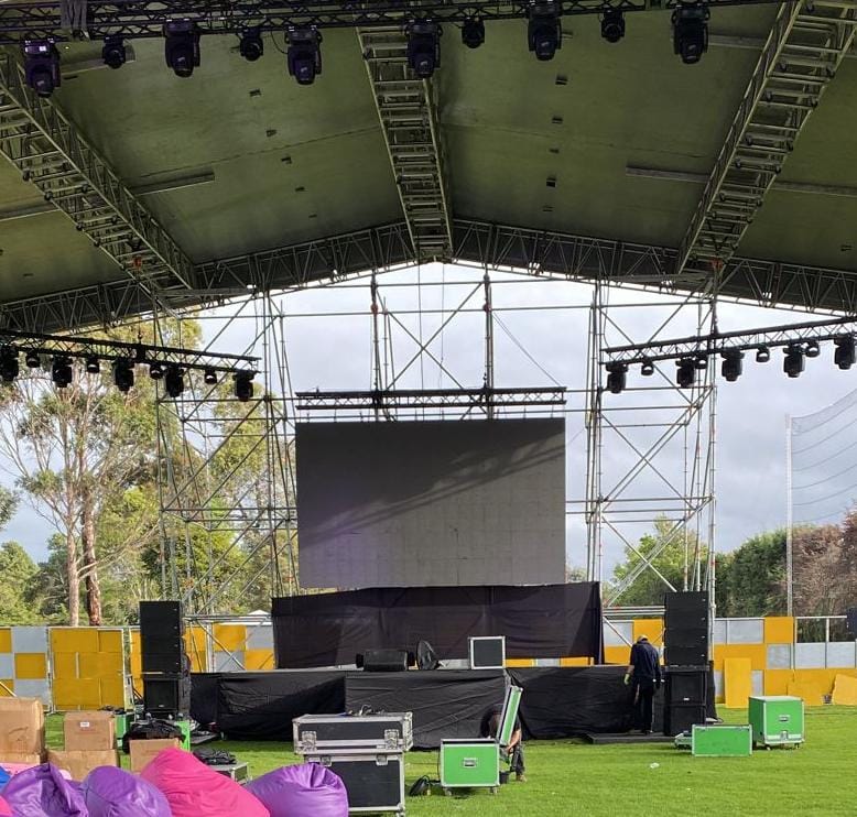 A covered outdoor stage with a large screen, lighting rig, speakers, and production equipment on a lawn