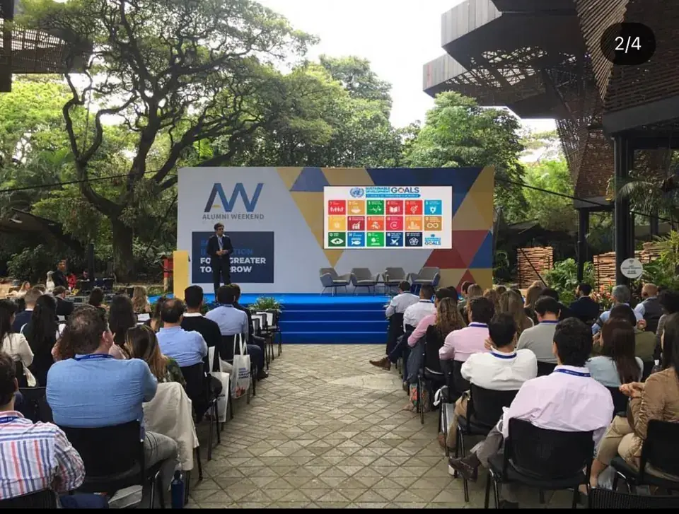 An audience seated outdoors beneath trees facing a large presentation screen